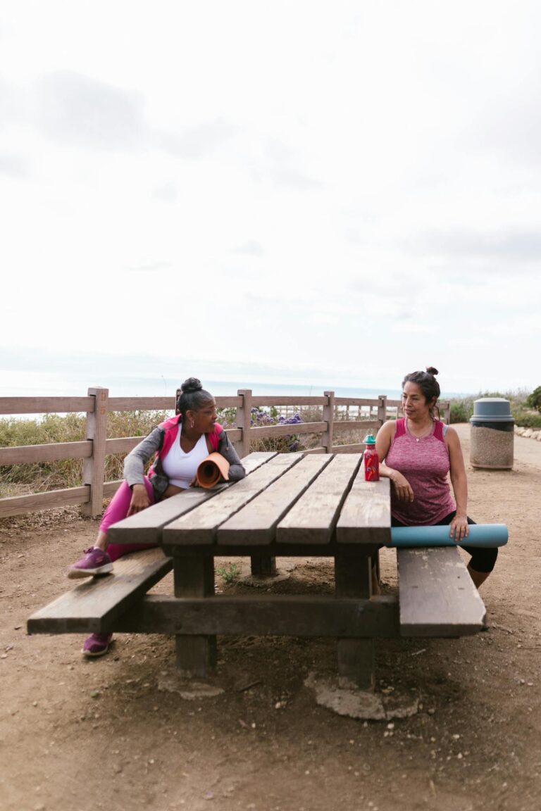 Two women take a break from yoga practice at a scenic seaside picnic area, enjoying the outdoors.