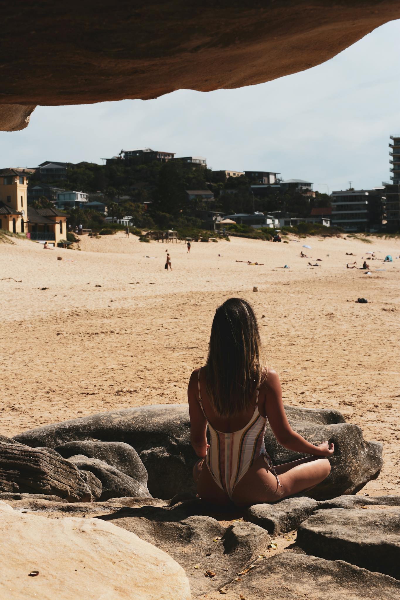 Woman in swimsuit meditating on a rock with a beach view under summer skies.